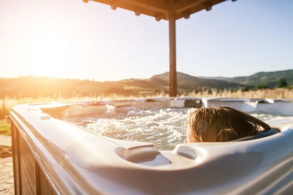 A woman relaxes in an outdoor hot tub and watches the sunset in the distance