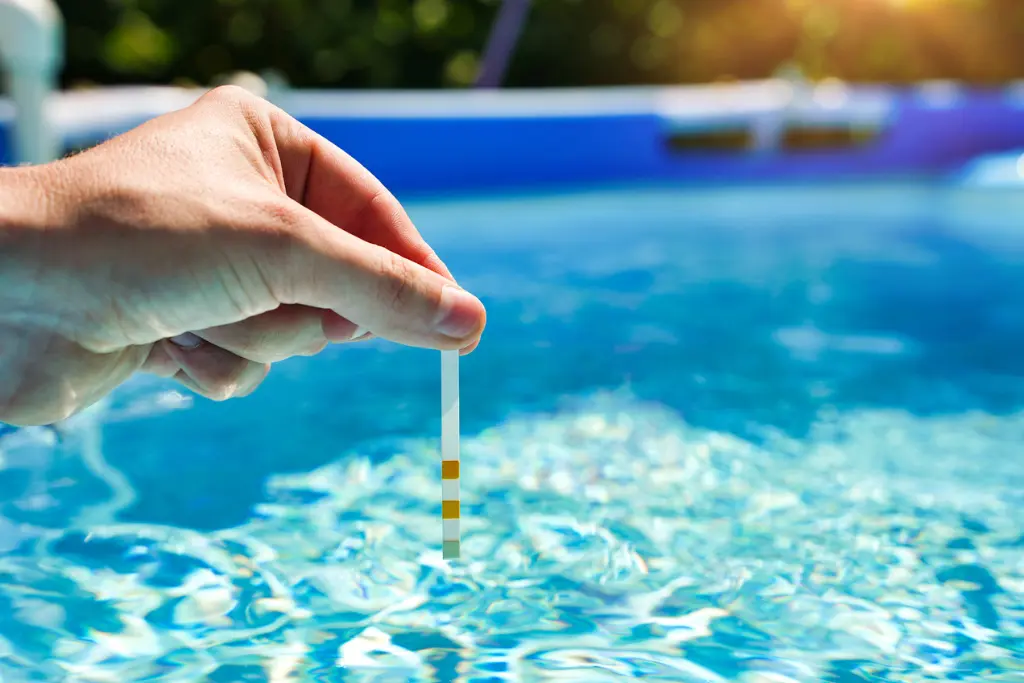 A man's hand dips a test strip into an above=ground pool's water.