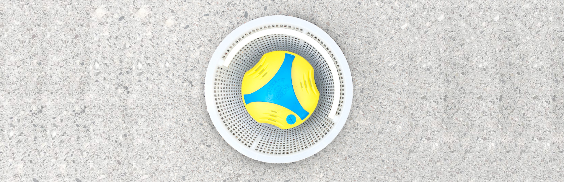 A top-down view of a yellow and blue pool chlorine dispenser sitting in a white basket strainer against a light gray concrete or stone surface. The circular device has a blue center with yellow curved elements.