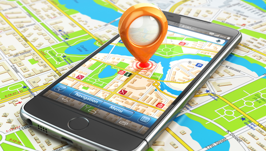 Smartphone displaying GPS navigation map with orange location pin marker, placed on top of colorful printed street maps