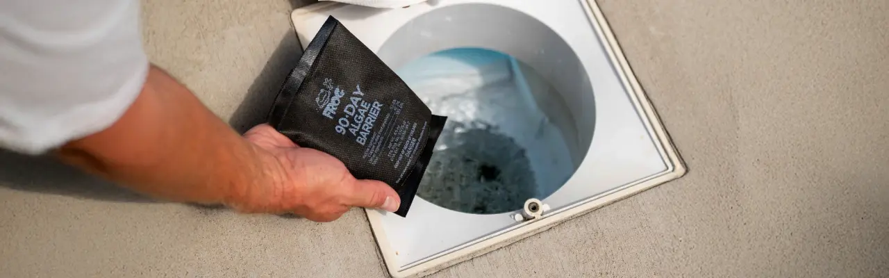 A person holds a black packet labeled '90 DAY ALGAE BARRIER' above an open pool skimmer with the lid removed, showing water and debris inside. The packet is a product intended to prevent algae growth in swimming pools.