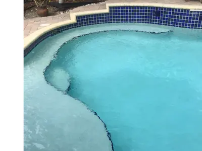 A swimming pool with cloudy blue water and a curved step area leading into the deeper section. The pool is bordered by a tiled edge and a paved patio, with potted plants, trees, and outdoor furniture including a chair and table in the background.