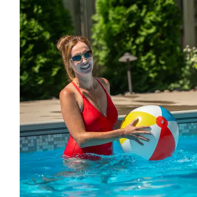 A person in a red swimsuit stands in a swimming pool holding a colorful beach ball. Green bushes and outdoor furniture are visible in the background.